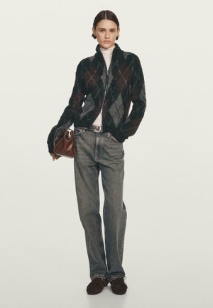 Czarny i szary kardigan z zamkiem błyskawicznym w wzór argyle, biały golf, wyblakłe niebieskie dżinsy, trzymając brązową torbę o zakrzywionym wzorze.