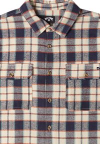 Camisa de cuadros abotonada hecha de franela. La combinación de colores incluye azul marino, blanco, naranja y verde azulado. Dos bolsillos en el pecho y botones marrones.