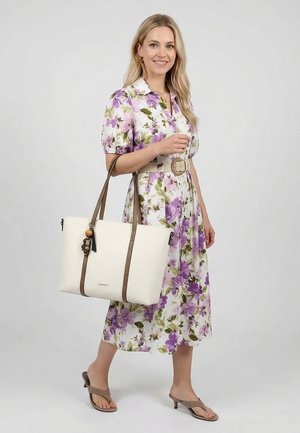 Femme souriante, portant une robe midi à fleurs violettes avec une ceinture, des sandales à talons beiges, et portant un grand sac fourre-tout blanc avec des sangles marron.