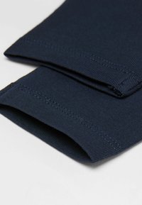 Detailní záběr na tmavě modrou tkaninu s precizním šitím na složených okrajích rukávů na světlém pozadí.