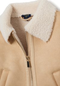 Veste en suède beige avec un col en fourrure crème, fermeture éclair dorée et poches à rabat. Texture douce, design décontracté adapté aux enfants.