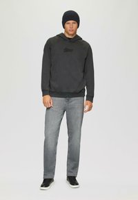 Czarna bluza z kapturem, z prążkowanym dołem, szare dżinsy i czarne sneakersy. Model nosi ciemną czapkę beanie, stojąc na jednolitym tle.