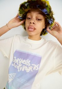 Crème kleurig T-shirt met een grafische print in blauw en paars, voorzien van de tekst "Schelpjes & Surfplanken." Fluffy, veelkleurige bucket hat.