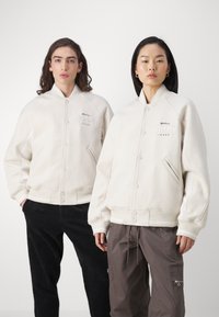 Svetlá krémová bomber bunda s rebrovaným golierom a manžetami, s predným zapínaním na gombíky a vyšitým logom "TOMMY JEANS" na hrudi.