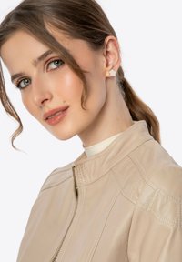 Beige leren jack met staande kraag en zichtbare stiksels. Het model heeft golvend haar en draagt bloemenoorbellen.