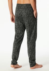 Barefoot person wearing dark green pajama pants with an all-over black leaf pattern, standing with one hand in pocket.