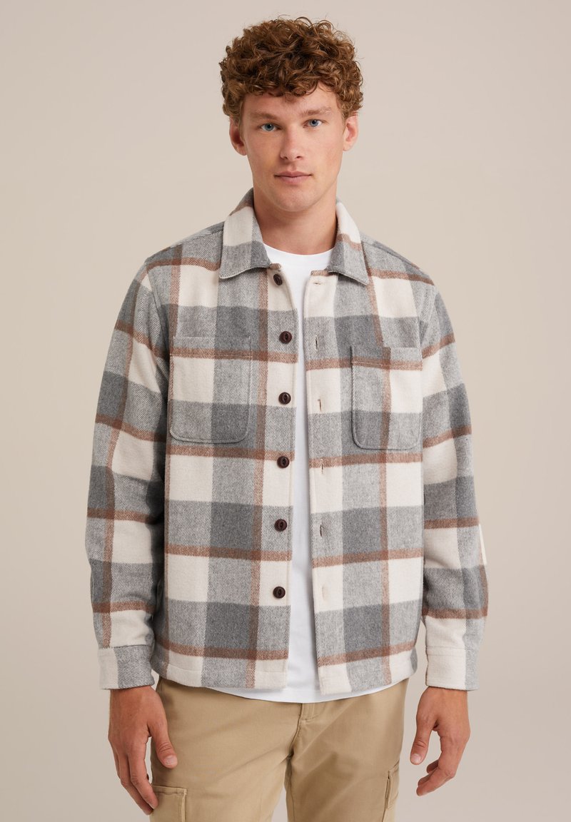 Młody mężczyzna ubrany w brązową, szaro-białą w kratkę koszulę z guzikami nałożoną na białą koszulkę oraz beżowe spodnie, stojący na jednolitym tle.