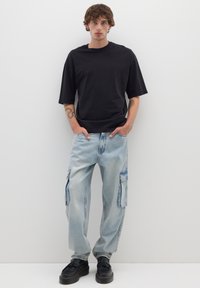 T-shirt nera a maniche corte abbinata a jeans cargo blu chiaro con tasche laterali e scarpe con suola piatta nere con superficie testurizzata.