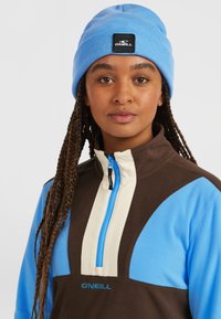 Blauwe beanie en een ritsfleece-top met bruine en lichtblauwe panelen en een logo. De stof lijkt zacht en textuur te hebben.