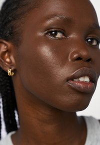 Zbliżenie twarzy o gładkiej, ciemnej skórze, delikatnym blasku i lekkiej teksturze. Cechy to warkocze oraz mała złota kolczyk w kształcie obręczy.