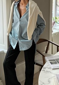 Camicia azzurro chiaro a maniche lunghe con bottoni, sovrapposta a un maglione beige. Pantaloni neri su misura. Tessuti morbidi, vestibilità rilassata e stile casual.