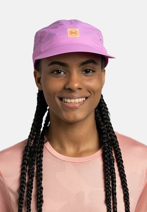 Smiling person with long braids wearing a pink breathable shirt and matching pink cap with an orange Buff logo on the front.