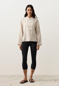 Femme portant une chemise beige à col mandarin avec des boutons en forme de grenouille, des leggings noirs courts et des sandales noires, debout sur un sol en pierre.