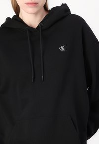 Felpa nera con cappuccio realizzata in un tessuto misto cotone, con maniche lunghe, cordini e un piccolo logo bianco "cK" sul petto.