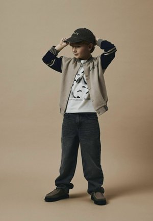 Chaqueta de varsity gris con mangas azul marino, camiseta blanca con estampado, jeans azules y zapatos grises. Un niño ajusta una gorra oscura. Fondo neutral.