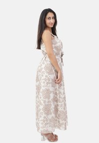 Kvinde med langt mørkt hår iført en ærmeløs beige blomster maxi kjole og transparente hæle, poserer sidelæns på en hvid baggrund.