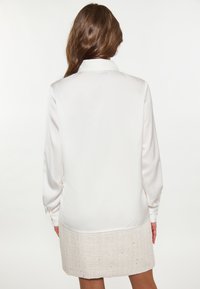 Valkoinen satiininen pitkähiha blouse klassisella kauluksella, yhdistettynä teksturoituun beigeen mini hameeseen, jossa on koristeellisia yksityiskohtia.