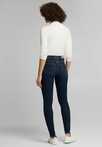 Vaqueros skinny de cintura alta en denim azul oscuro, con bolsillos traseros y costuras naranjas en contraste, combinados con una blusa blanca de manga larga.