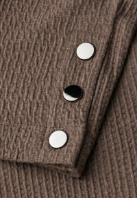 Tessuto a coste marrone con superficie texture; tre bottoni chiusura in argento. Finitura pulita con dettagli di cucitura sottili.
