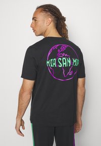 Camiseta de algodón negra con un gráfico colorido de un globo en la parte trasera con "MIA SAN MIA" en letras verdes y moradas. Mangas cortas, corte casual.
