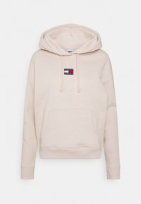 Beige mikina z měkké látky, která má klokani kapsu a nášivku s logem na hrudi. Obsahuje kapuci se šňůrkou a žebrované manžety.
