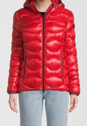Personne portant une doudoune matelassée rouge brillante avec une capuche, une fermeture éclair noire et deux poches zippées, associée à un jean bleu clair.