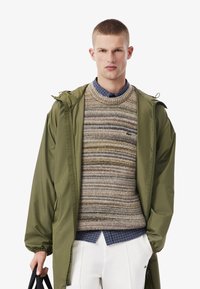 Giacca leggera verde oliva con cappuccio e maniche arricciate, indossata sopra un maglione di lana a strisce, stratificato su una camicia a quadri blu.