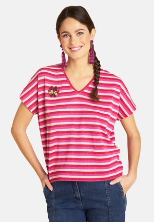 Woman with braided hair smiling, wearing pink and red striped V-neck shirt with embroidered hand patch, pink tassel earrings, and blue jeans.