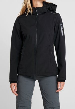 Frau trägt eine schwarze Jacke mit Reißverschluss und Kapuze sowie einer Reißverschlusstasche am oberen linken Ärmel, kombiniert mit grauer Hose.