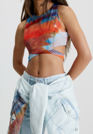 Frau trägt ein mehrfarbiges, gemustertes Crop-Top mit seitlichen Ausschnitten und hellblaue Jeans mit einer Jeansjacke, die um die Taille gebunden ist.