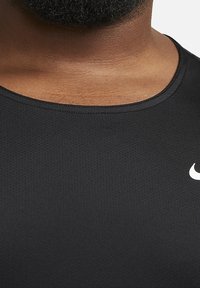 Chemise de sport noire en tissu texturé et évacuant l'humidité. Présente un col rond et un logo blanc sur la poitrine.