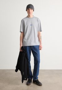 Graues Baumwoll-T-Shirt mit kleinem blauen Logo, kombiniert mit blauen Jeans und einer schwarzen Jacke. Das Model trägt eine graue Strickmütze.
