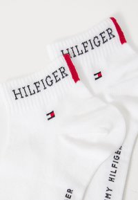 Hvide ankelstrømper med sort "HILFIGER" tekst på kanten, røde detaljer og et lille Tommy Hilfiger-logo nær ankelen. Blød strikket tekstur.