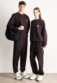 Tmavohnedé mikiny a tepláky vyrobené z mäkkého materiálu s malými geometrickými logami. Čierna taška a biele tenisky dopĺňajú outfity.