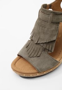 Sandale femme à bout ouvert en daim taupe avec détails à franges superposées, semelle intérieure rembourrée et bordure cloutée le long du bord de la semelle.
