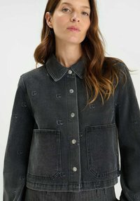 Mujer con cabello largo castaño que lleva una chaqueta gris oscuro abotonada con grandes bolsillos frontales y sutiles patrones bordados.
