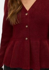 Bordo pletena jopica z V-izrezom, tremi velikimi rjavimi gumbi in peplum robom. Mehka tekstura z rebrastim vzorcem po celotni površini.
