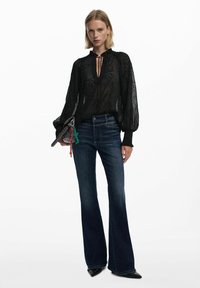 Blusa nera trasparente con ricami floreali e un collo alto, abbinata a jeans a campana blu scuro e scarpe nere a punta.