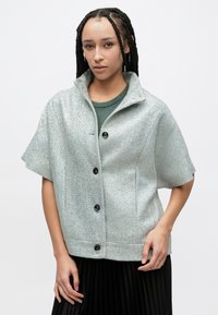 Lyseblå herringbone kortærmet jakke med krave, med sorte knapper og en afslappet pasform, båret over en grøn top.