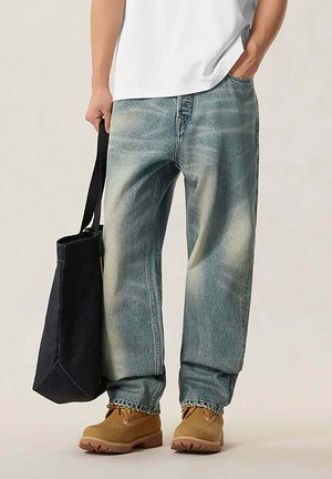 Persona indossa jeans blu sbiaditi e larghi, stivali da lavoro color cuoio e una camicia bianca, tenendo una grande borsa nera al fianco.