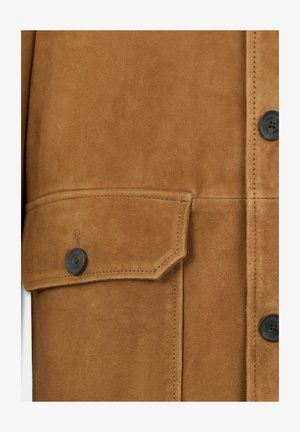 Veste en suède marron avec une poche à rabat boutonnée, des boutons noirs et des détails de couture visibles. Texture lisse avec une coupe ajustée.