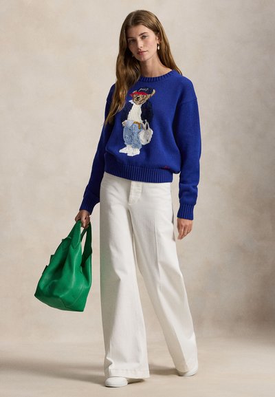 Πλεκτό πουλόβερ σε βασιλικό μπλε με κεντητό σχέδιο αρκούδας, συνδυασμένο με λευκό παντελόνι φαρδιάς γραμμής και πράσινη τσάντα χειρός, πάνω σε ουδέτερο φόντο.