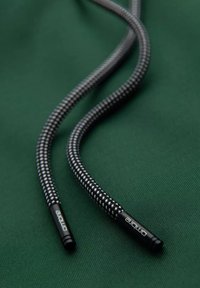 Svart flätad snodd med en slät yta, med metalländar märkta med "ombre", framför en mörkgrön bakgrund.