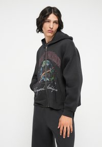 Felpa nera con zip e una stampa grafica colorata della Terra accompagnata da un testo. Presenta una pratica tasca a marsupio e polsini a coste. Tessuto morbido al tatto.