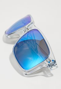 Ochelari de soare mari și transparenți, cu lentile gradiente de culoare albastră și accesorii din metal argintiu. Temele opuse prezintă mici accente albastre.