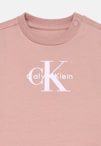 Gaiši rozā kokvilnas krekls ar apaļu kakla izgriezumu, plecu aizdari un pārāk lielu baltu "CK" logotipu ar tekstu "Calvin Klein".