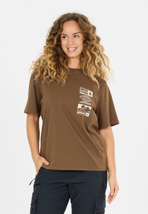 Lächelnde Frau mit lockigem Haar trägt ein braunes T-Shirt mit Grafik und schwarze Cargohosen, steht mit einer Hand in der Tasche vor weißem Hintergrund.