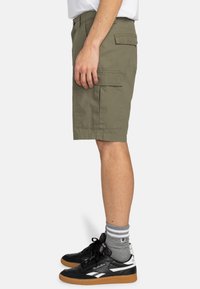Shorts de carga en verde oliva con bolsillos laterales, trabillas y un corte recto. Combinados con zapatillas negras y calcetines a rayas grises.