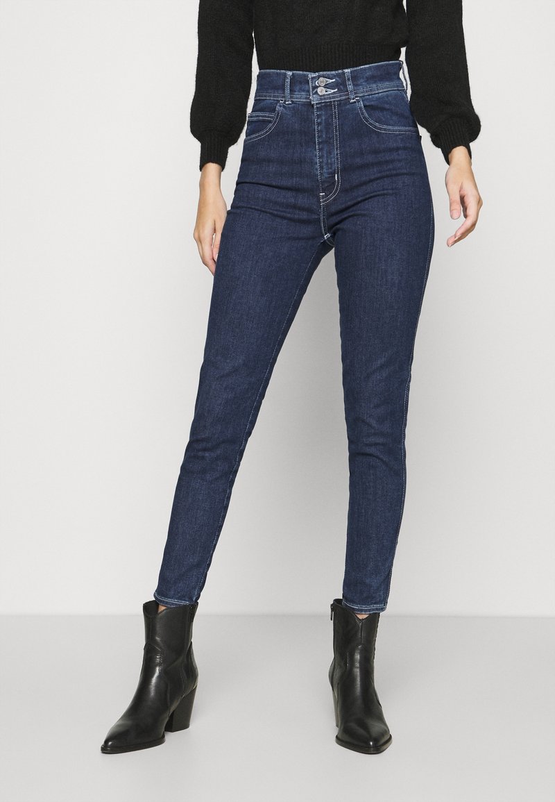 Jean skinny taille haute bleu marine avec un design classique à cinq poches et des accents de couture, associés à des bottines noires.