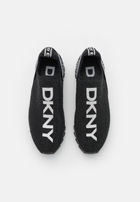 Pár černých tenisek typu slip-on s bílým logem DKNY na vrchu, texturovanou látkou a značkovými poutky na patách.